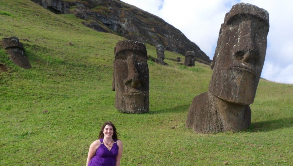 a woman in purple dress sitting in front of large carved stone statues with Easter Island in the background