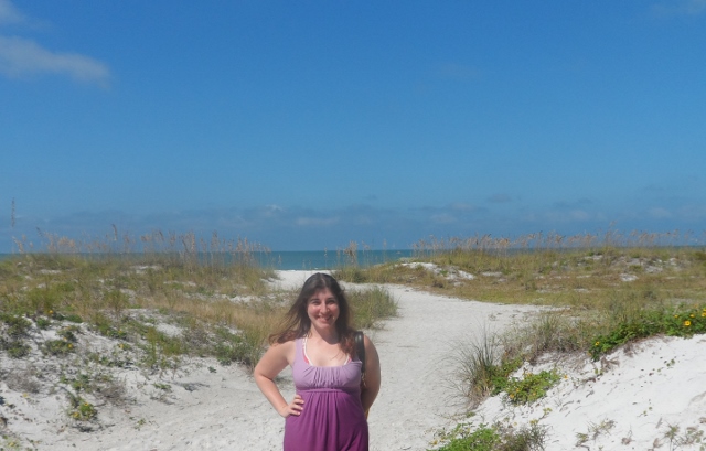 a woman standing on a sandy path with grass and blue sky