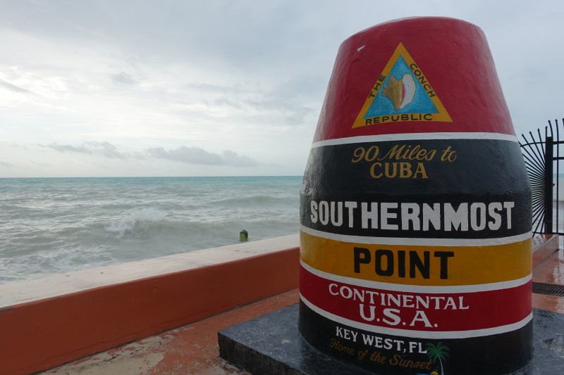 a sign on a stone wall with Southernmost point buoy in the background